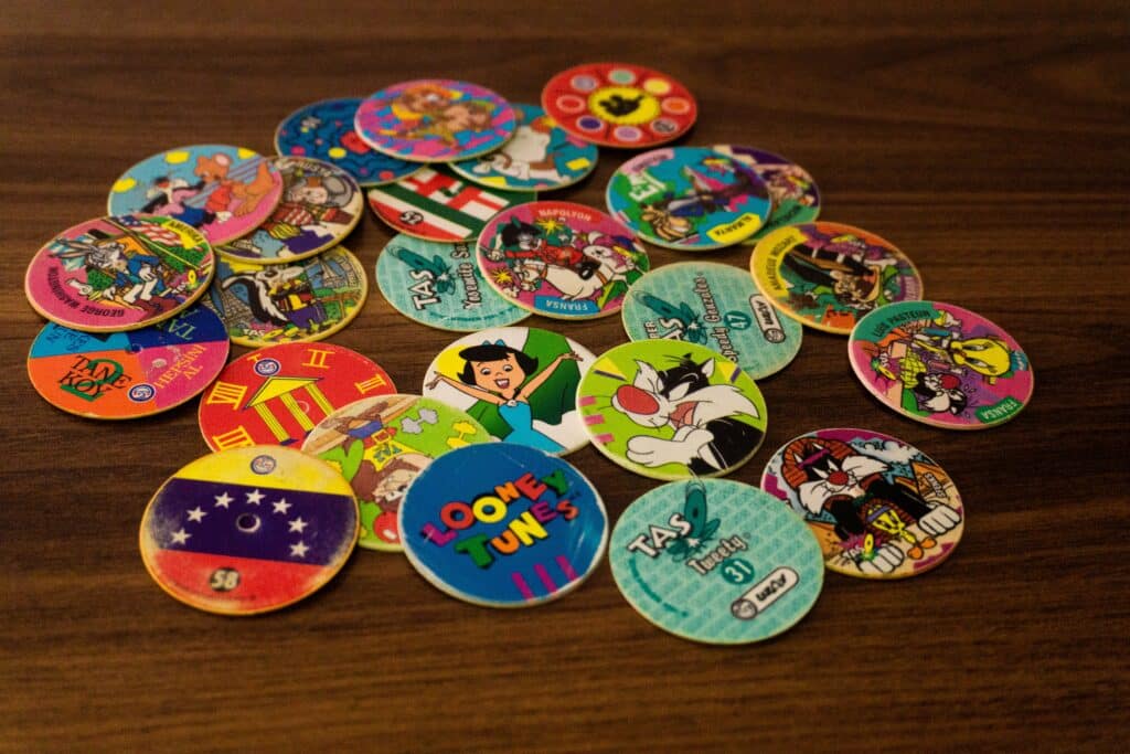 A scattered pile of colorful collectible game tokens, known as Pogs, featuring various cartoon characters such as Looney Tunes and The Flintstones, on a wooden surface. The tokens are round and made of cardboard or plastic, with vibrant illustrations and logos on each.