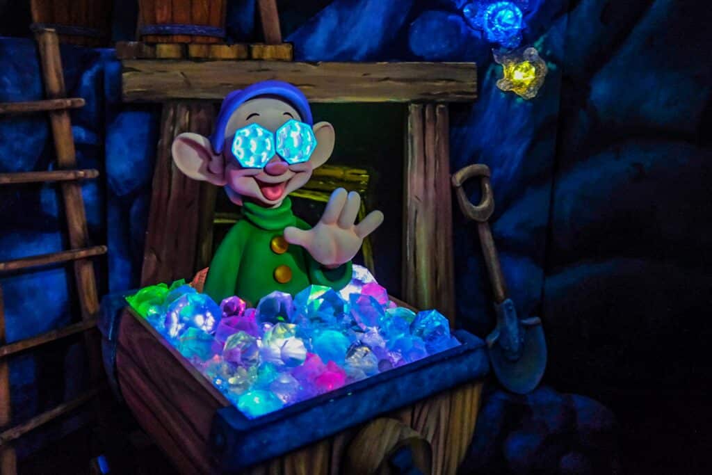 A colorful, animated scene featuring a cheerful dwarf character from Snow White, wearing large, glowing blue gemstone glasses and a green outfit with yellow buttons. The dwarf is waving from inside a wooden mining cart overflowing with brightly lit, multicolored gems. The background depicts a cave with glowing crystals embedded in the rock walls, a wooden ladder, and a shovel resting nearby.